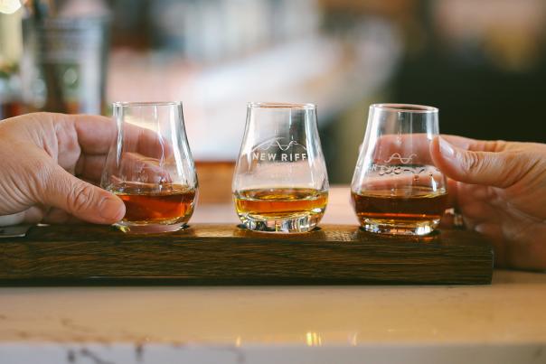 Three glencairn glasses, each with a pour of whiskey, sit in a wooden holder. Hands from either side reach out to select a glass.