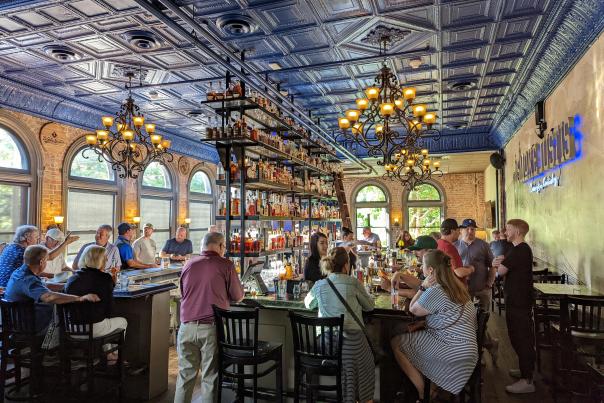 The bourbon bar at Smoke Justis with six shelves of bourbon in the center, surrounded by a wooden bar seated with many customers