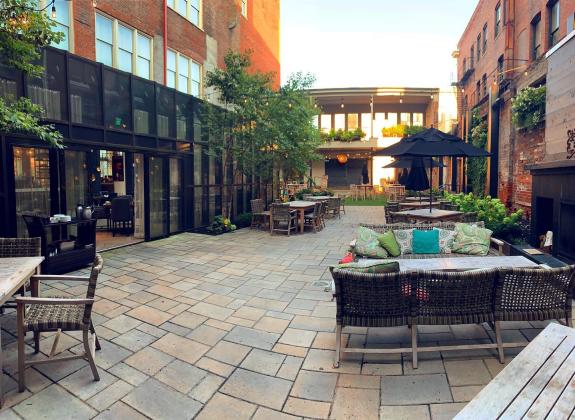 An outdoor patio, lush with plants, offers seating. A fireplace is to the right and an area that can be used as a stage is in the background.