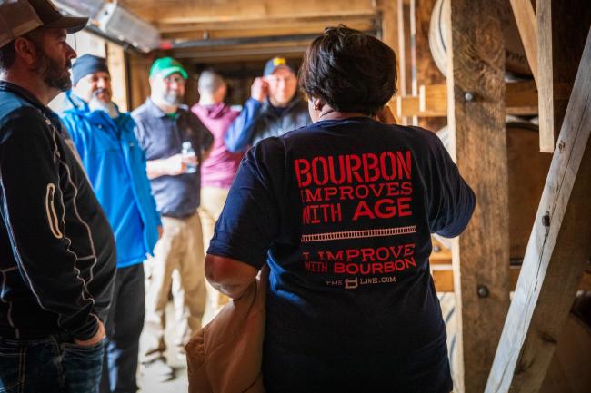 A woman faces away from the camera, speaking to a tour group. Her shirt says "Bourbon improves with age, but I improve with bourbon".