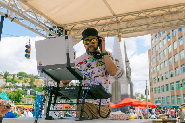 A DJ in a colorful shirt and sunglasses stands behind his equipment in the foreground, with a cityscape and a large monument visible in the background.