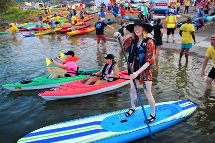 A woman in a hat and life vest stands on a paddleboard in the foreground, while kayaks and a crowd of people are visible in the water and on the shore in the background.
