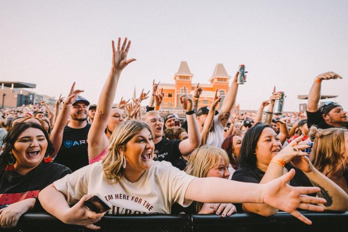 A crowd of excited people with raised hands and smiles are gathered in front of a stage at MegaCorp Pavilion in Newport, with a building visible in the background.
