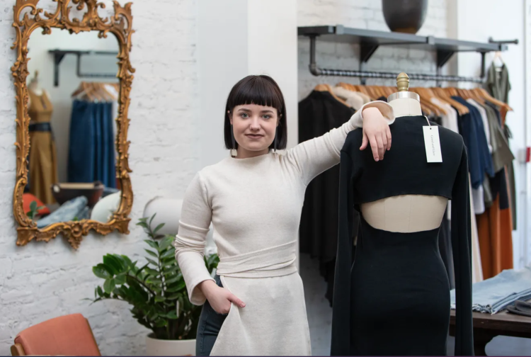 A woman faces the camera, she rests her arm on a mannequin and there are racks of clothing for sale behind her.