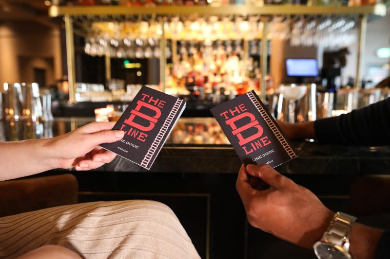 Two people's hands are are in the foreground, holding paper B-Line Guides. In the background is a bar with many bottles of bourbon.