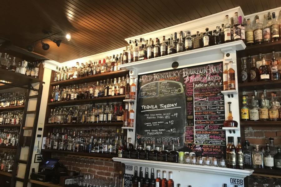 A wall covered in shelves of hundreds of bottles of bourbon, with a chalkboard menu in the middle at Old Kentucky Bourbon Bar