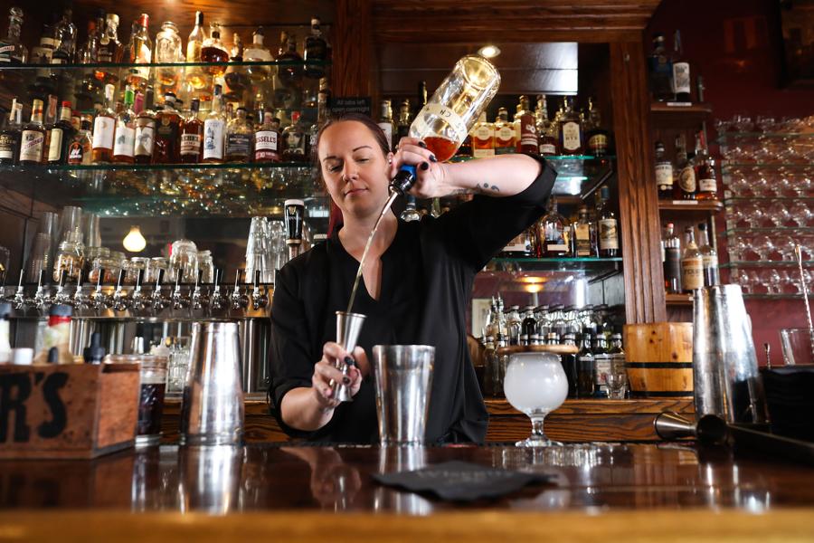 A bartender measures ingredients for a cocktail. In front of them is a shaker and behind are shelves of bourbons.