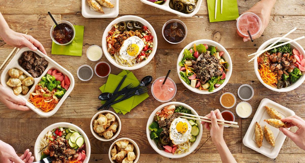 The image is of multiple white bowls and plates on a wood table with a variety of cuisine in them. The image also contains drinks and sauces.