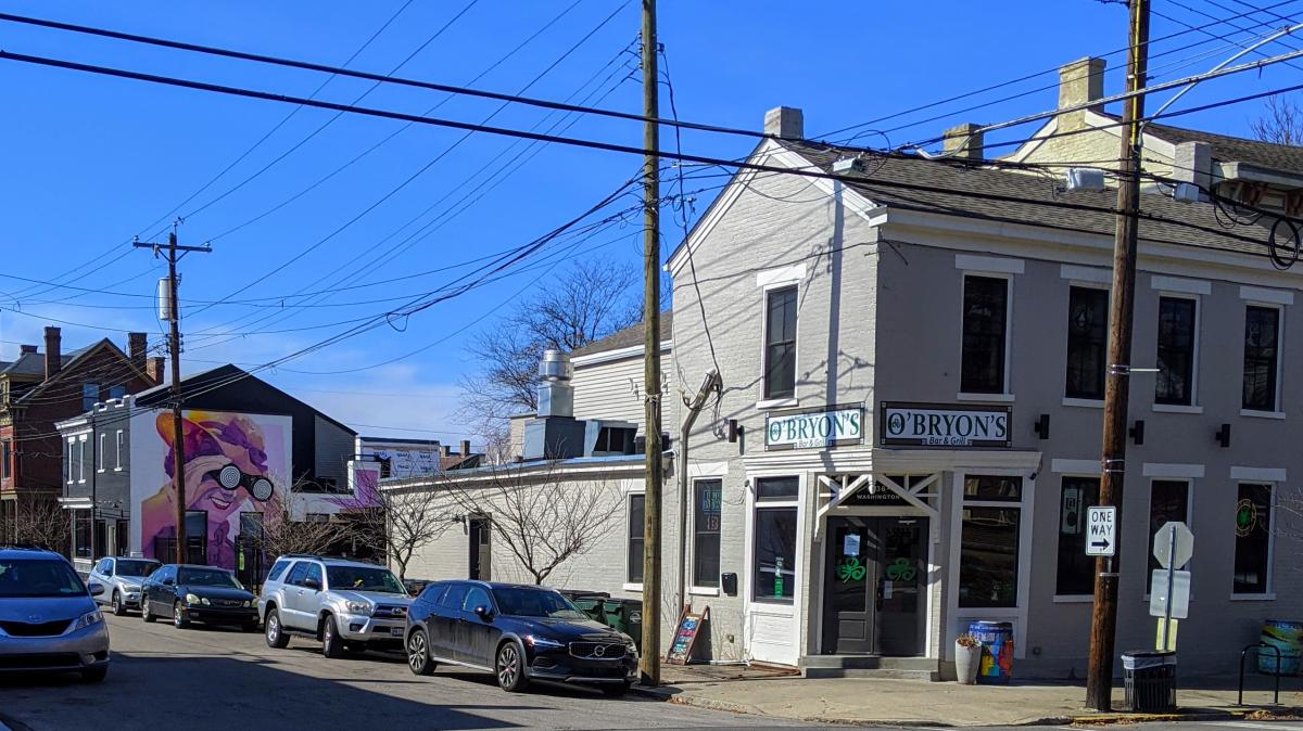 A row of gray buildings under a blue sky. One building has a mural of a lady holding binoculars. Another building on the corner has a sign that says O'Bryon's