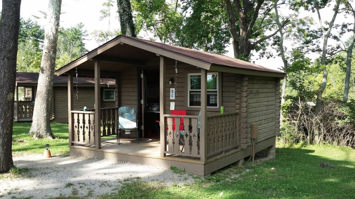 Image is of a small, one bedroom cabin next to the trees with a small porch in the front.