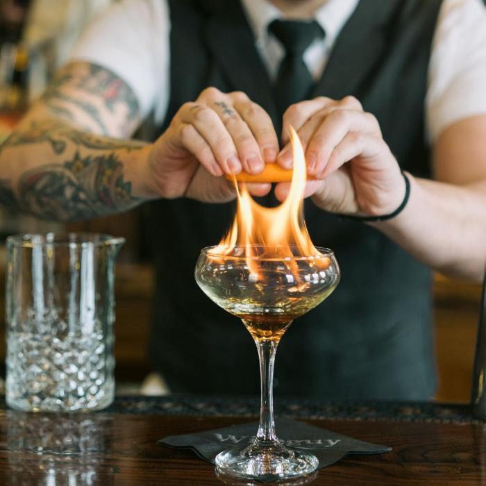 A bartender squeezes orange oil over a flaming cocktail.