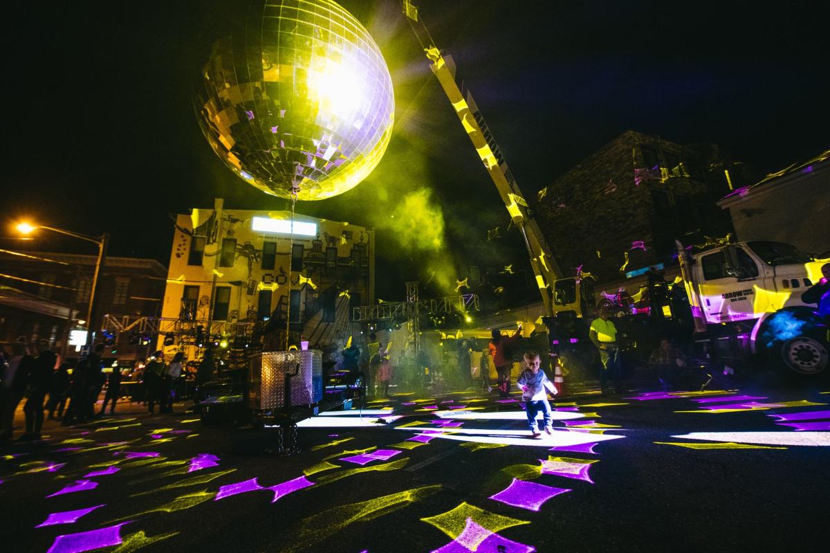 A giant disco ball hanging over a parking lot lighted up yellow with a baby dancing underneath