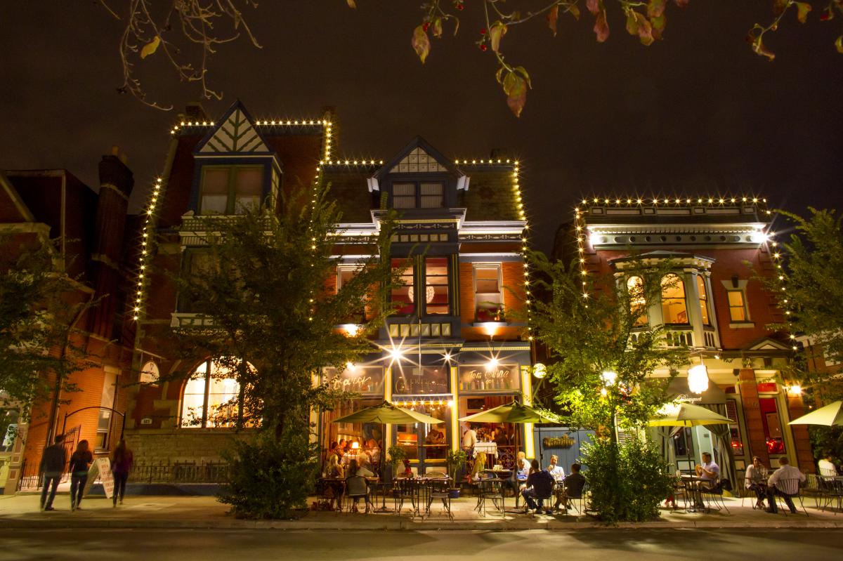 Three three story buildings lit up with strings of lights with diners eating at tables outside in front at night