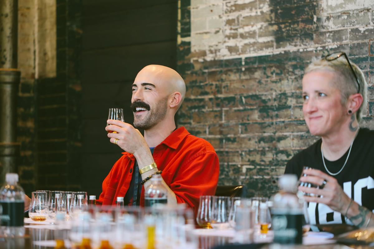 A male and female sitting against a brink wall tasting bourbon from a barrel pick.