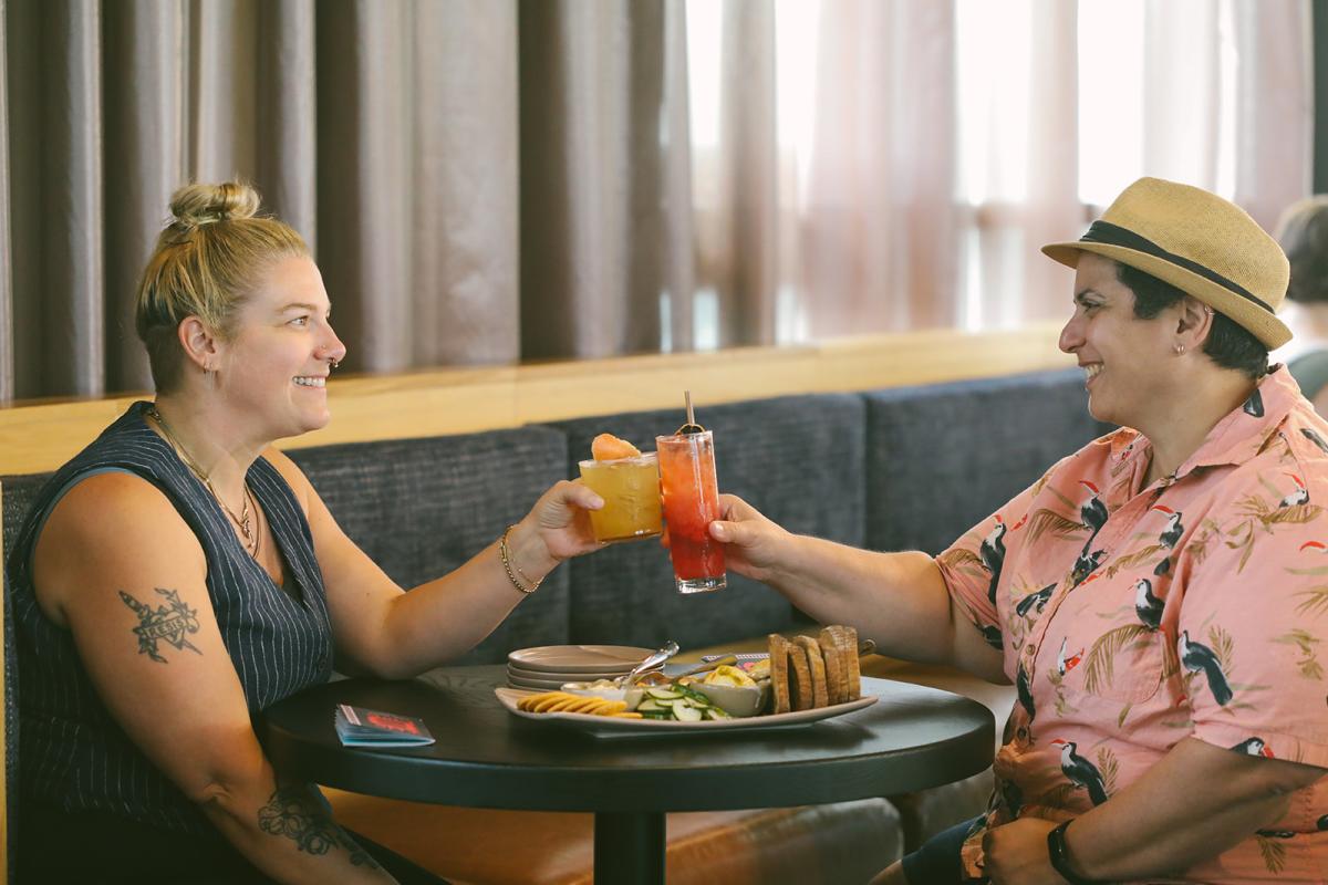 Two women toast each other with cocktails at a round table at the Aquifer bar at New Riff Distilling