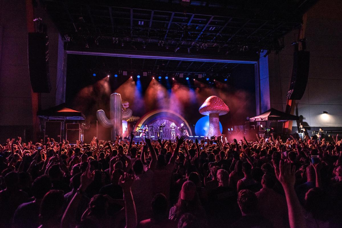 A stage bathed in purple and red light, with a giant mushroom and Kesha with a crowd of fans in the foreground.