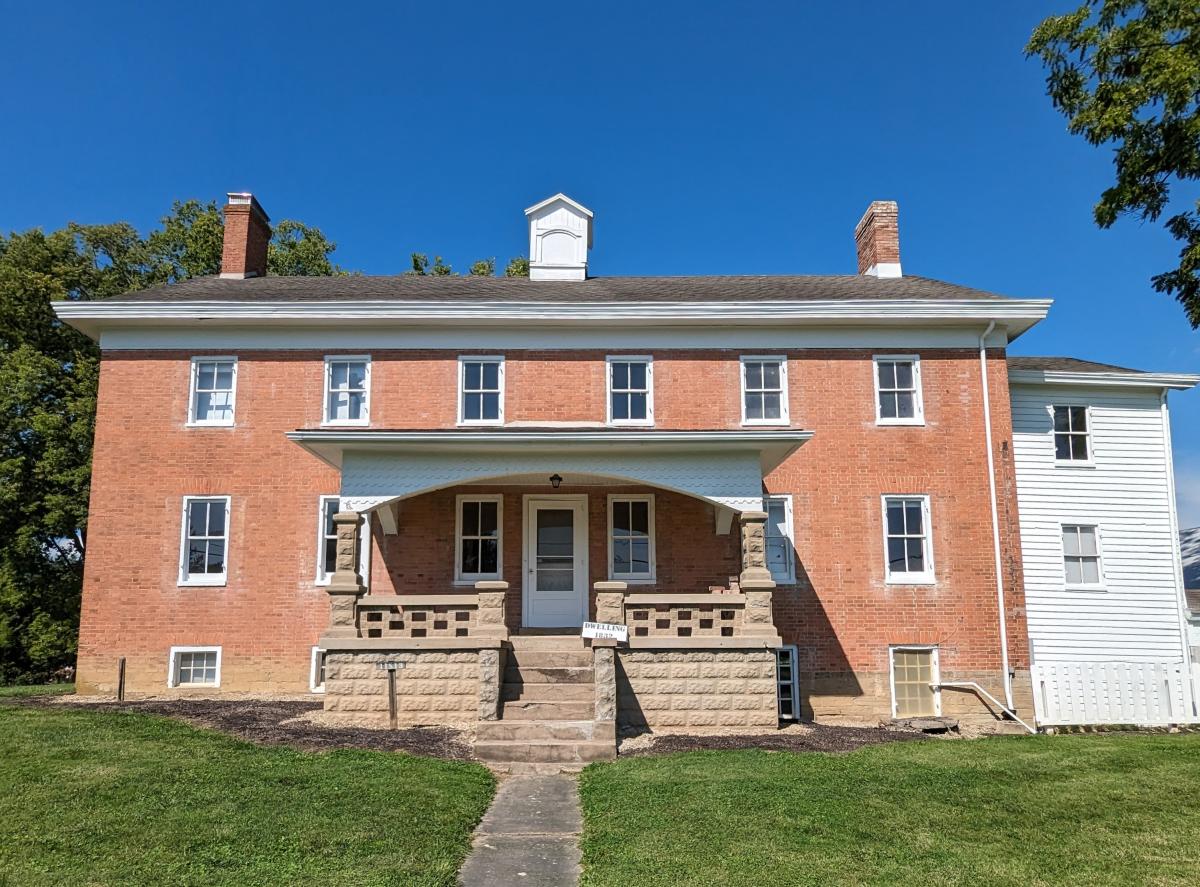 Image of the Dwelling House which is a two story, red brick house with a front porch and one door in the center.