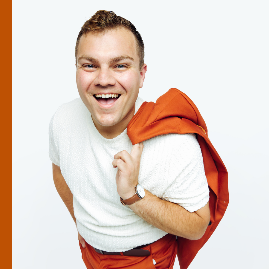 A smiling man in a white shirt and orange pants holds an orange jacket over his shoulder against a plain white background.