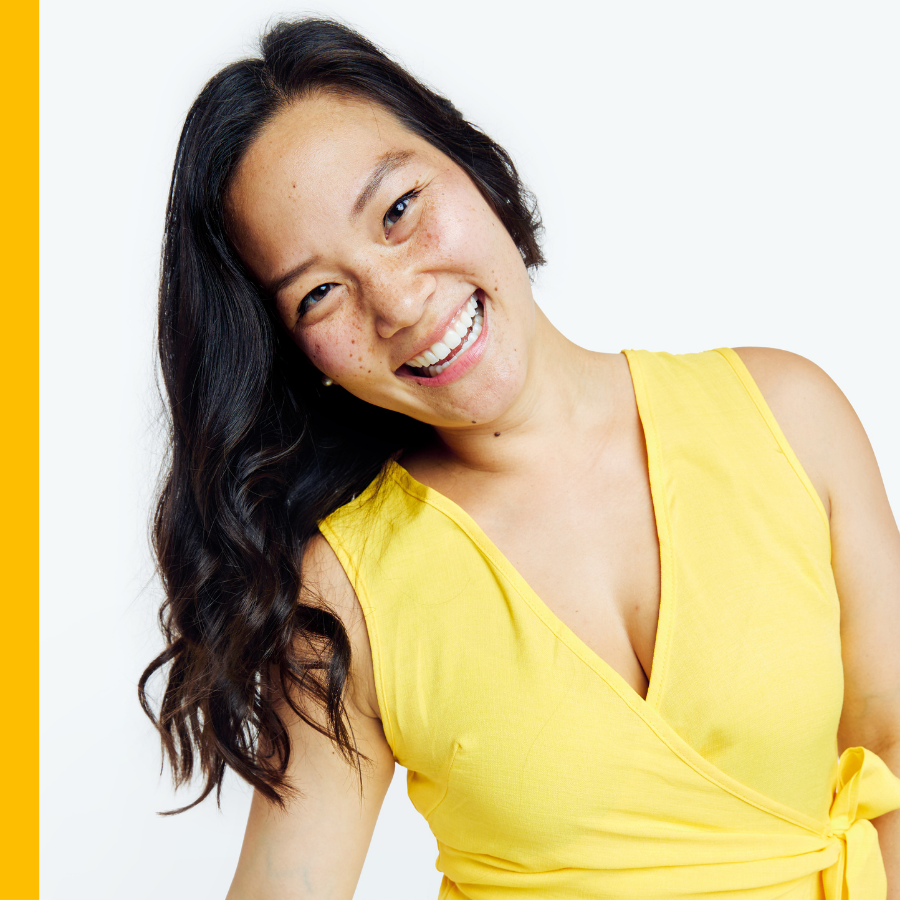 A smiling woman with dark hair and freckles in a yellow dress is against a plain white background.