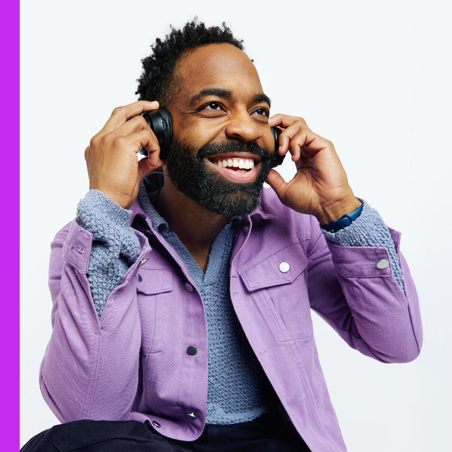 A smiling Black man in a purple jacket and grey sweater adjusts his headphones against a plain white background.