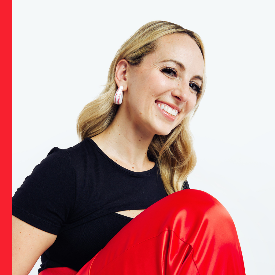 A smiling woman with blonde hair and large shell earrings, wearing a black top and shiny red pants, is captured against a plain white background.