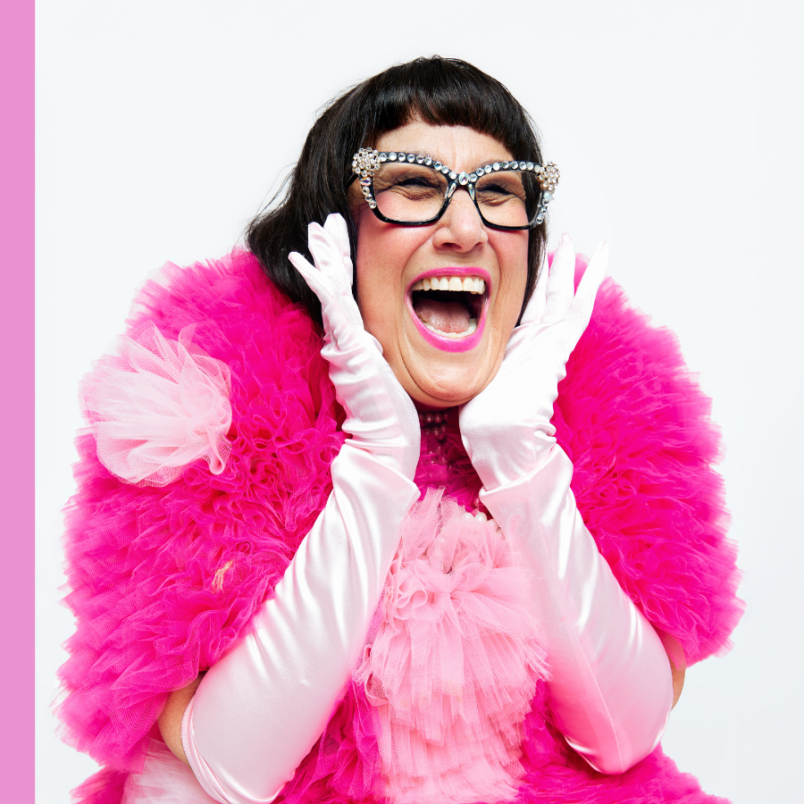 A woman in a vibrant pink fluffy outfit and long white gloves with her hands on her cheeks, her mouth open in a joyful expression, is set against a plain white background.
