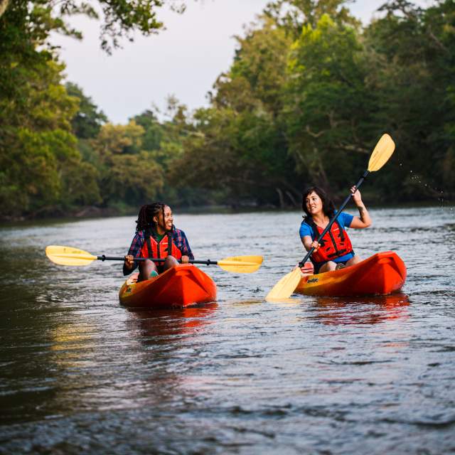 Two women paddle in a river in two orange kayaks.