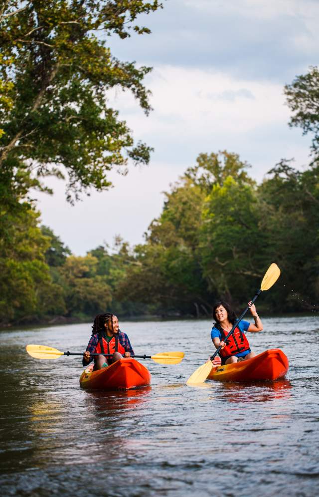 Two women paddle in a river in two orange kayaks.