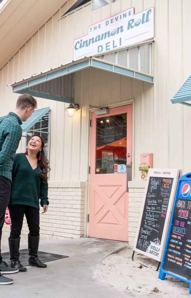 A couple is outside the entrance to the Devine Cinnamon Roll Deli about to enter.