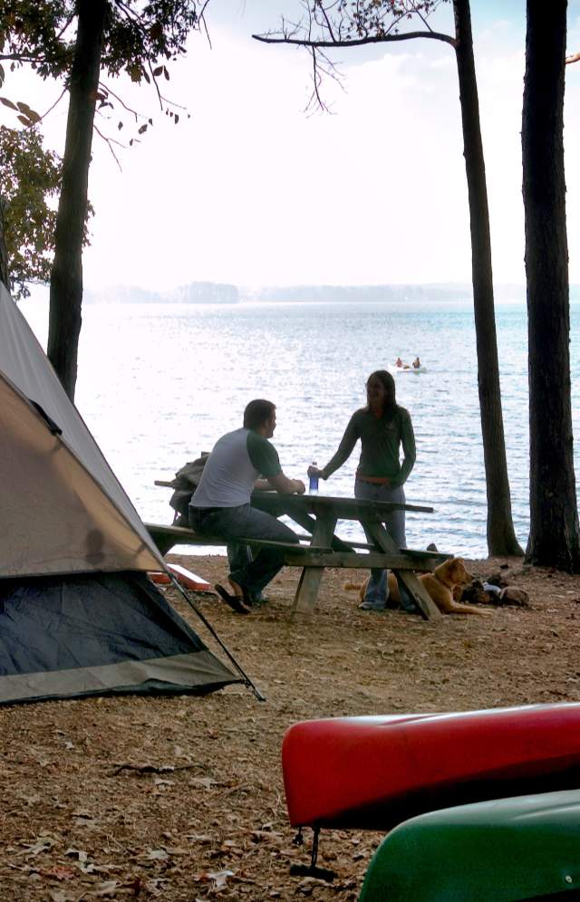 A couple with their dog on the shore of a lake next to the tent and kayaks