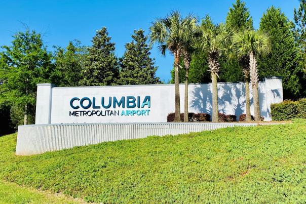 A large, grey, cement brick sign stands on top of a small hill. There are letters in blue that say "Columbia Metropolitan Airport" on the left side with 5 Palmetto trees on the right side.