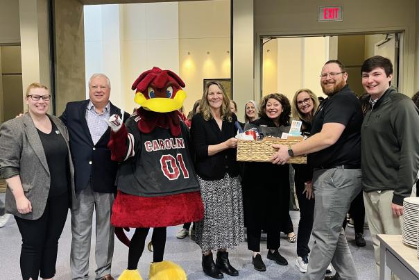 A group of people pose together with Cocky the USC Gamecocks mascot. A basket is presented to someone in the middle in honor of the 4-millionth visitor to the convention center.