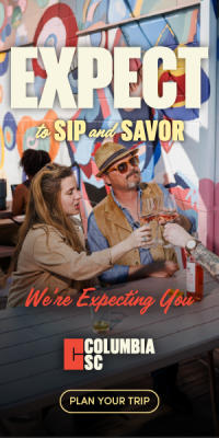 A couple is holding wine glasses filled with rose at a picnic table. They are holding the glasses together in the middle for a cheers.