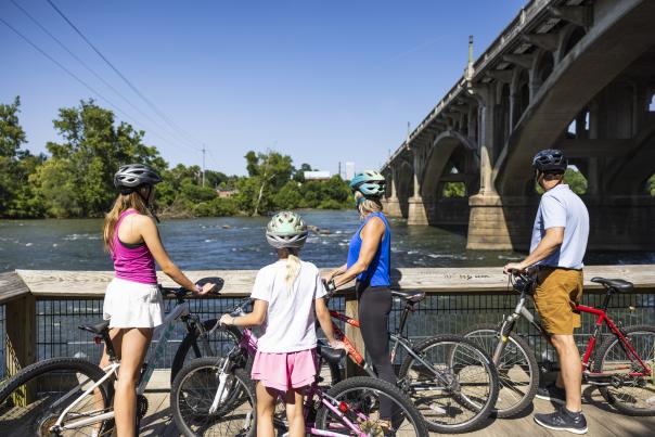 A family rides their bikes along the trail next to the river. There is a bridge in the background connecting one side of the river to the other.