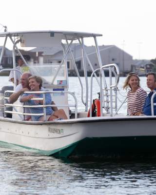 A small boat with four people glides across calm waters, surrounded by waterfront homes and trees.