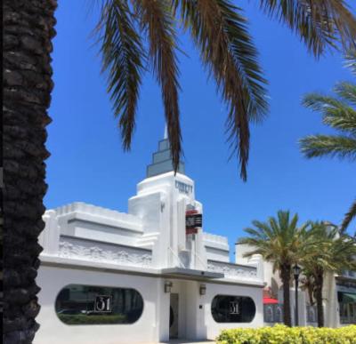 White building with a tall stepped tower, ornate scalloped trim, and rounded corner windows, framed by palm trees against a clear blue sky.