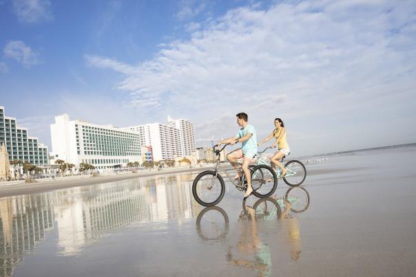 Two people riding bikes on the beach with tall buildings in the distance
