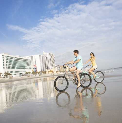 Two people riding bikes on the beach with tall buildings in the distance