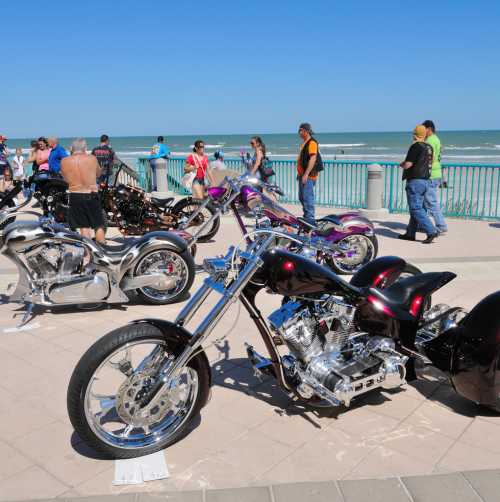 Several motorcycles parked along the boardwalk with a crowd around them