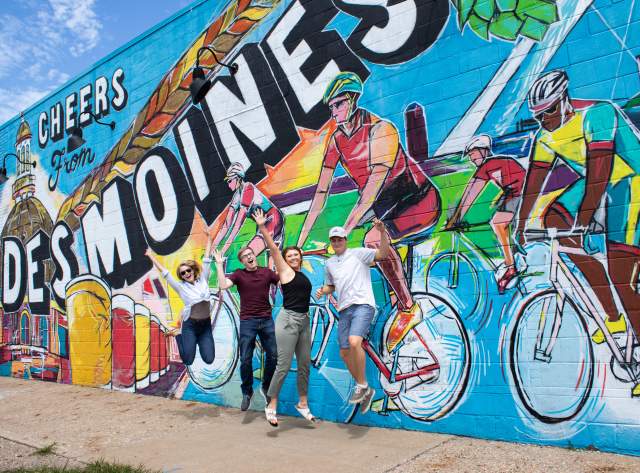 Friends jump in joy in front of the "Cheers from Des Moines" mural in downtown Des Moines.