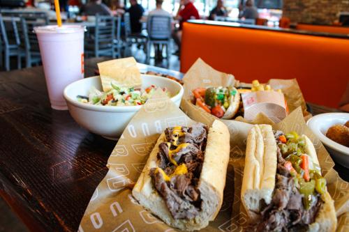 two Italian beef sandwiches, one with cheese and one with giardiniera. There is also a sandwich and a shake in the background.