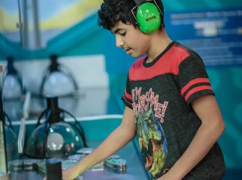 A boy wearing green noise-canceling headphones and a dinosaur T-shirt plays an interactive science exhibit, pressing glowing buttons on a table.