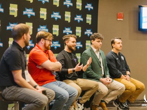 A panel of five men sit on stools during a discussion at the Midwest Battleground event, hosted by DuPage Sports. They are seated in front of a branded step-and-repeat backdrop. One of the panelists, dressed in a black sweatshirt, is animatedly speaking while the others listen. The atmosphere is professional yet casual, and the event appears to focus on esports or gaming, as indicated by the logos and attire.