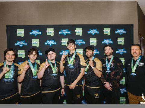 A victorious esports team poses in front of a Midwest Battleground and DuPage Sports Commission step-and-repeat banner. The seven team members wear matching black and gold jerseys and proudly display their medals, with several holding up a finger to signify their first-place win. They are smiling confidently, celebrating their achievement at the gaming competition.