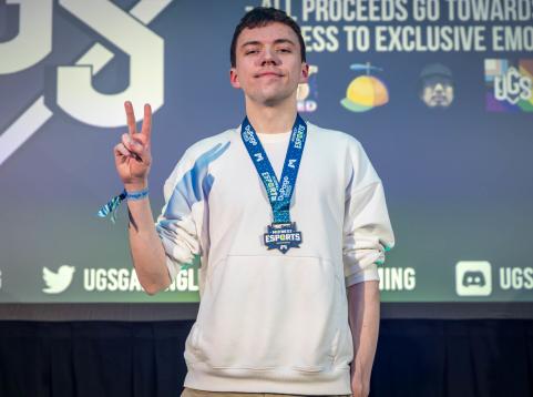 A young man stands on stage wearing a white sweatshirt and a 'Midwest Esports' medal around his neck. He holds up a peace sign with his right hand and smiles confidently at the camera. Behind him, a screen displays logos and social media handles related to the event, with the letters 'UGS' prominently visible. The scene suggests the conclusion of an esports competition.