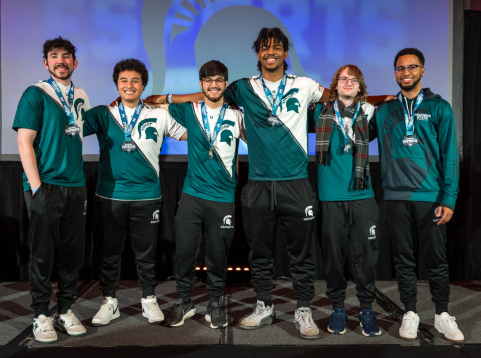 Six members of the Michigan State University esports team stand together on stage, wearing matching green and white jerseys with the Spartan logo. Each team member proudly displays a medal around their neck, smiling at the camera after an esports event. A large Spartan helmet logo is visible in the background, highlighting their university pride.