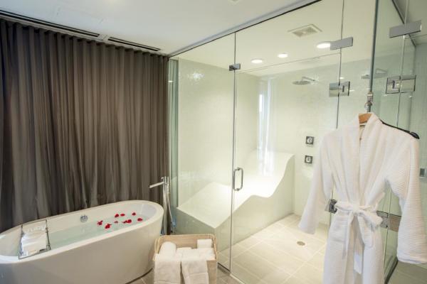 A modern spa bathroom featuring a glass-enclosed steam shower with a built-in seating area, a freestanding bathtub with rose petals, and a hanging white bathrobe. Towels are neatly folded in a basket nearby, and a dark curtain separates the space for privacy.