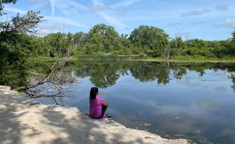 Woman in pink shirt sitting on rocks next to blue lake with green trees in background