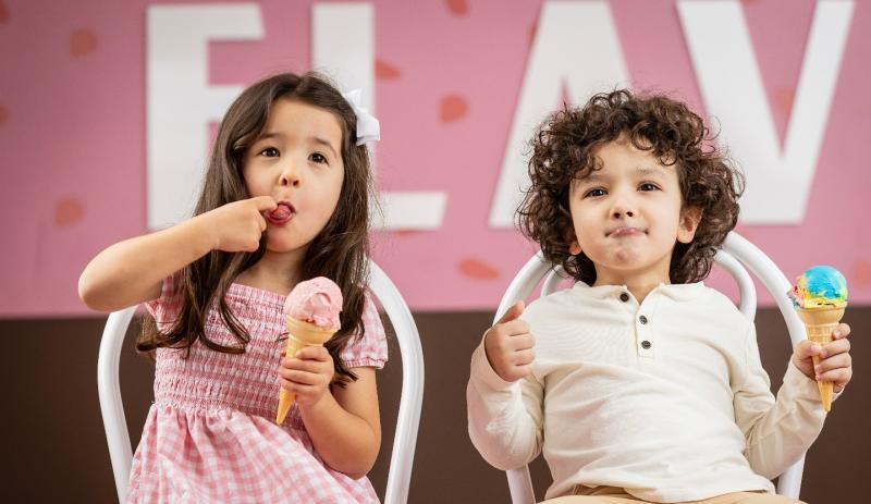 kids eating ice cream. There is a girl in a pink dress licking her pointer finger. She is eating strawberry ice cream. The boy sitting next to her is wearing a white shirt and is holding an ice cream cone in his left hand.