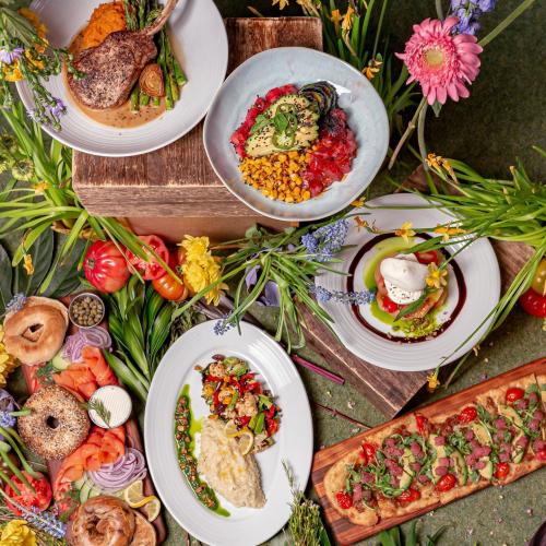 Colorful brunch spread with flatbread, lamb chops, risotto, bagels, and dessert, surrounded by fresh flowers.